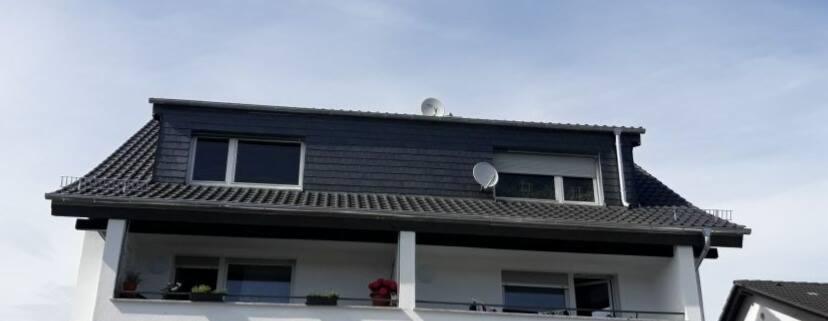 Dwupiętrowy dom z balkonem i dachem pokrytym ciemną dachówką, widoczne okna dachowe i dwie anteny satelitarne.