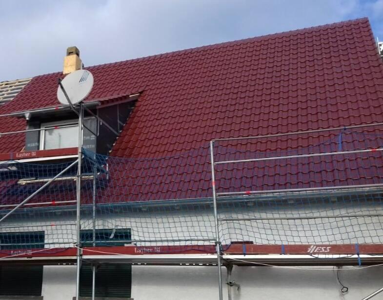 Czerwony dach z dachówki karpiówki, częściowo osłonięty siatką ochronną na rusztowaniu, widoczny komin i antena satelitarna.