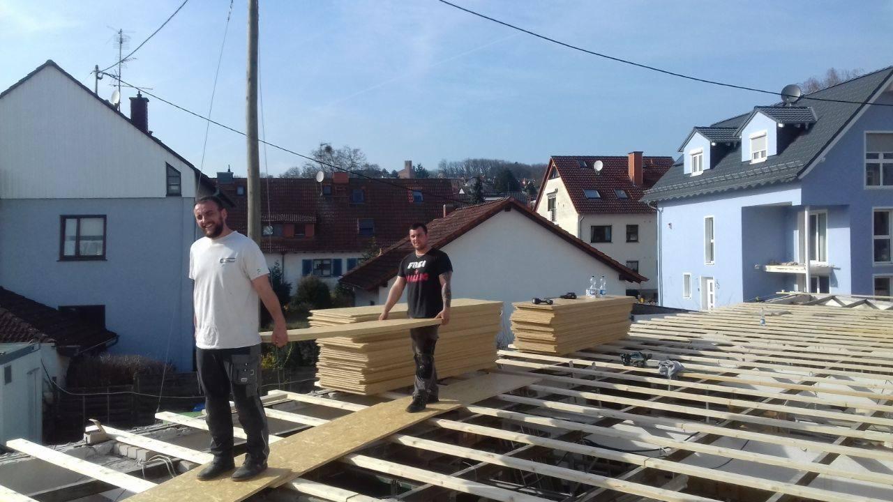 Dwóch mężczyzn układa płyty na drewnianej konstrukcji dachu, w tle zabudowa mieszkalna