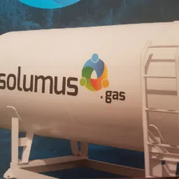 Biały zbiornik z logo 'solumus .gas' i drabinką, na tle niebieskiego tła z motywem lasu.