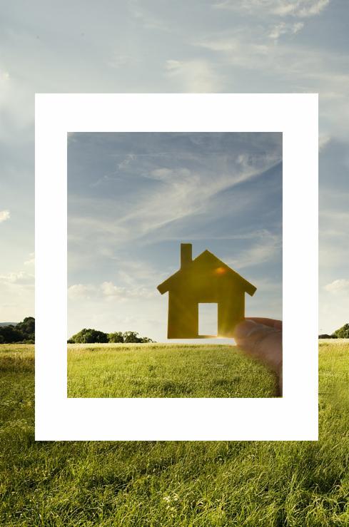 Dłoń trzyma wyciętą z papieru sylwetkę domu na tle zielonego pola i błękitnego nieba z chmurami, słońce prześwieca przez otwór drzwiowy.
