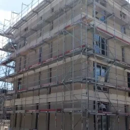 Wielopiętrowy budynek w trakcie nakładania jasnoszarej elewacji, otoczony rusztowaniami z widocznymi napisami 'Gerüstbau Steiner', z niezagospodarowanym terenem wokół.