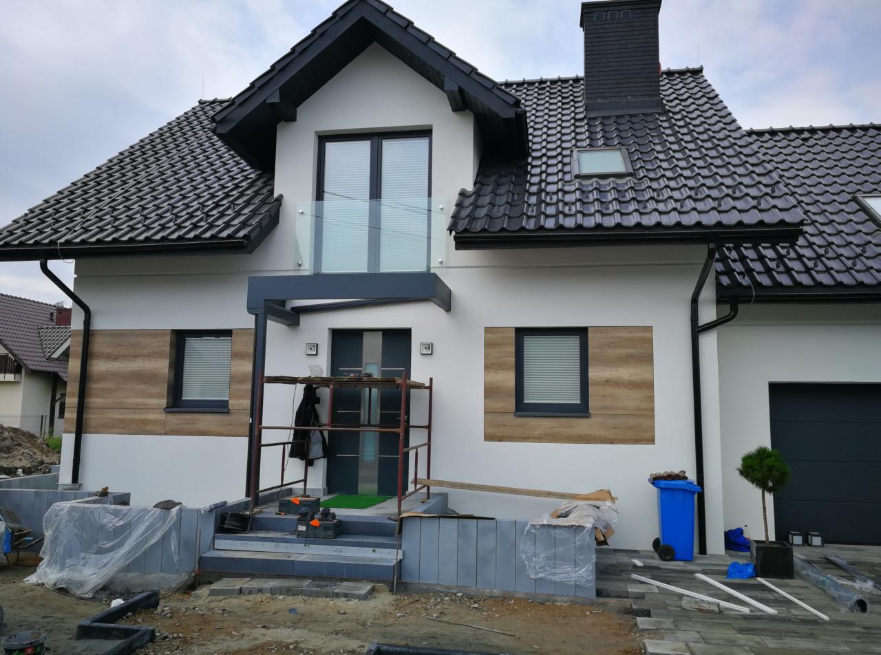 Nowoczesny dom z białą elewacją, elementami drewnopodobnymi i czarną dachówką, z widocznym rusztowaniem przed wejściem i trwającymi pracami brukarskimi.