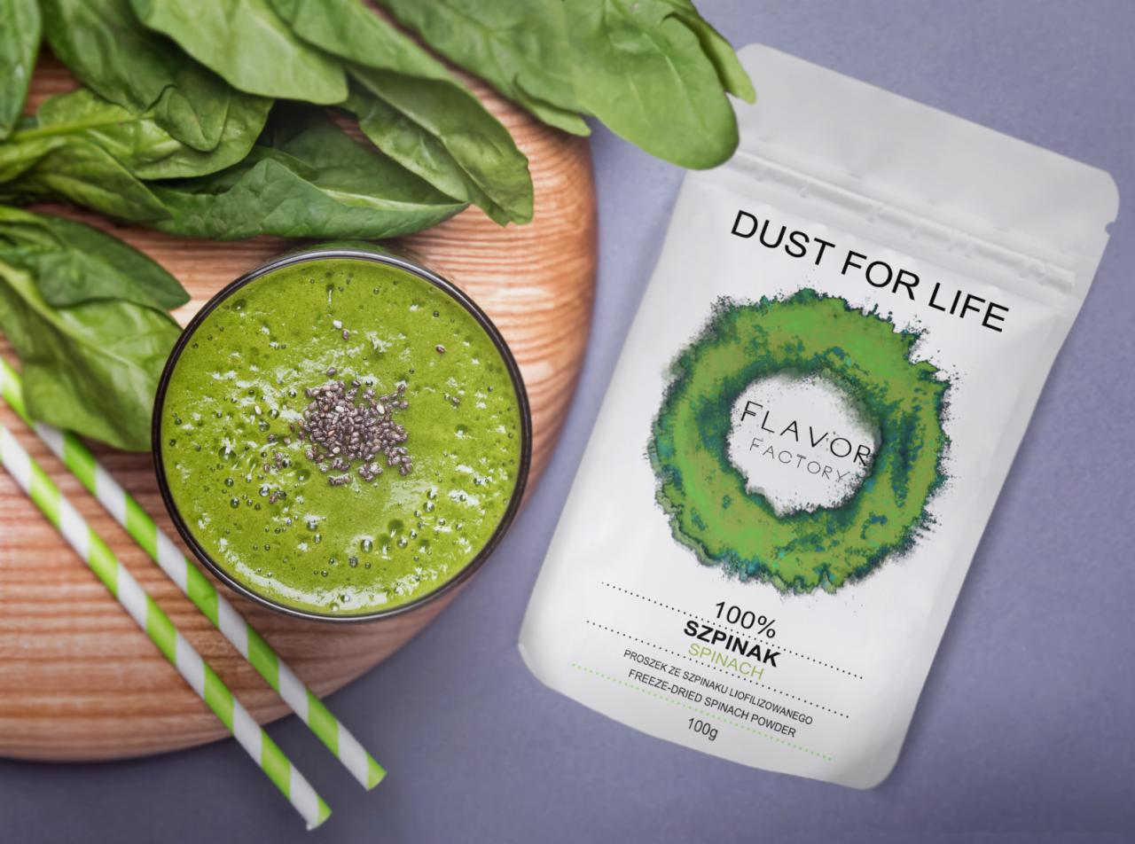 Zielony koktajl ze szpinakiem i nasionami chia w szklance, obok świeże liście szpinaku na drewnianej desce i opakowanie sproszkowanego szpinaku od Flavor Factory.