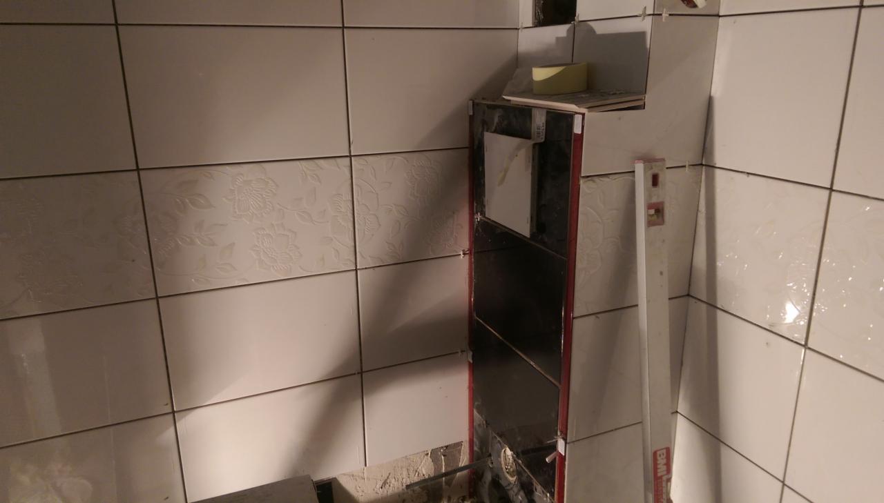 Fragment łazienki w trakcie układania białych płytek ściennych, częściowo z delikatnym kwiatowym wzorem, widoczny pion poziomicy i przygotowane miejsce na półki.