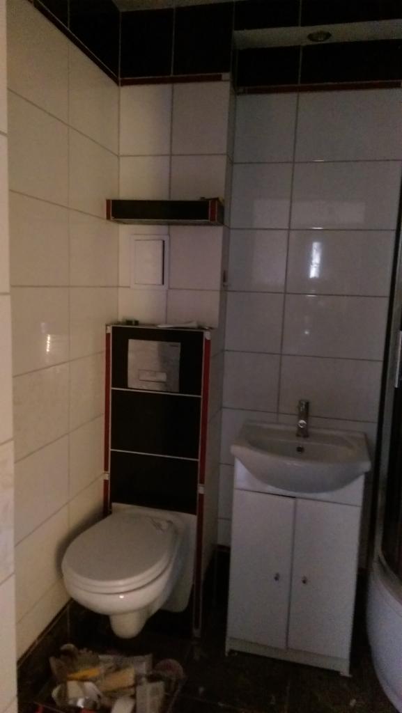 Wykończona łazienka z podwieszaną toaletą w zabudowie z czarnymi i czerwonymi akcentami, białe płytki na ścianach, biała umywalka z szafką, widoczne akcesoria do montażu na podłodze.