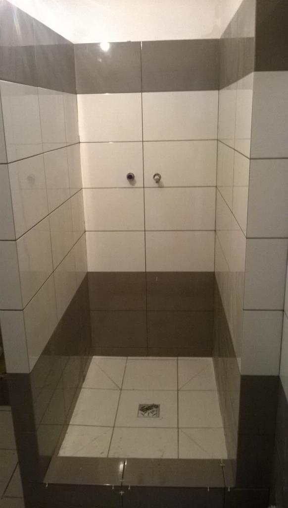 Wykończony brodzik z biało-szarymi płytkami ceramicznymi, widoczne dwa metalowe zawory na ścianie i odpływ liniowy w posadzce.