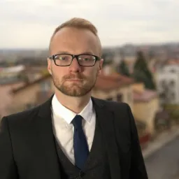 Portret mężczyzny w garniturze, z brodą i okularach, na rozmytym tle miejskiego krajobrazu z dachami budynków.