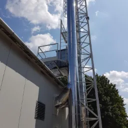 Błyszczący, metalowy komin przemysłowy z drabinką, przymocowany do ściany budynku na tle błękitnego nieba z chmurami.