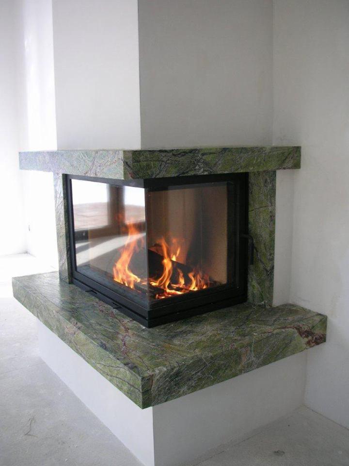 Nowoczesny kominek narożny z obudową z zielonego marmuru, płomienie widoczne za szybą.