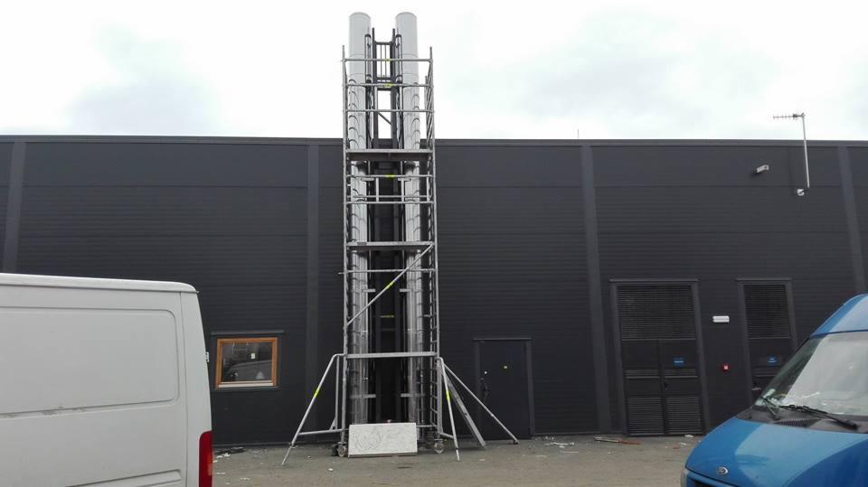 Dwa pionowe kanały wentylacyjne w trakcie montażu na zewnątrz budynku komercyjnego, umieszczone na wysokim rusztowaniu, widoczne dwa samochody dostawcze w tle.