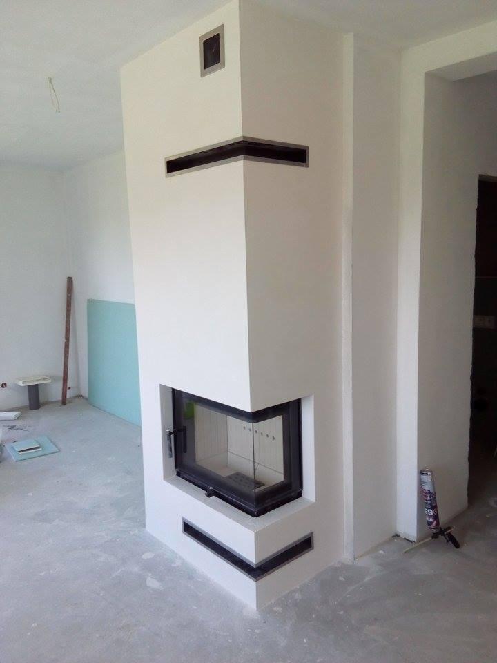 Biały, narożny wkład kominkowy z czarnymi detalami w surowym wnętrzu, widoczna tuba silikonu i płyta gipsowa w tle.