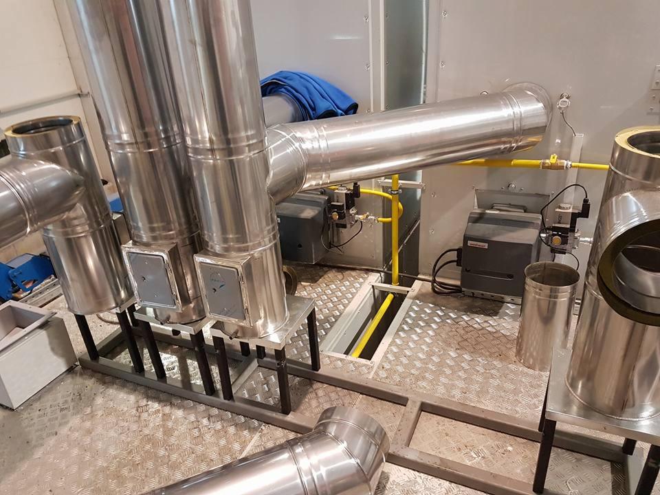 Instalacja z rur wentylacyjnych ze stali nierdzewnej, żółte rury gazowe i czarne urządzenia, widoczne na metalowej podłodze w pomieszczeniu technicznym.