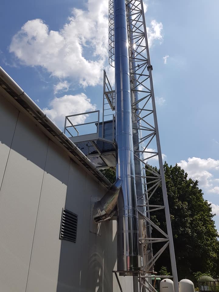 Błyszczący, metalowy komin przemysłowy z drabinką, przymocowany do ściany budynku na tle błękitnego nieba z chmurami.