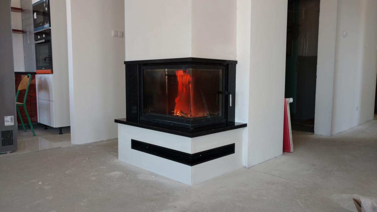 Kątowy kominek z czarną obudową i widocznym płomieniem, umieszczony w surowym wnętrzu z betonową podłogą i białymi ścianami.