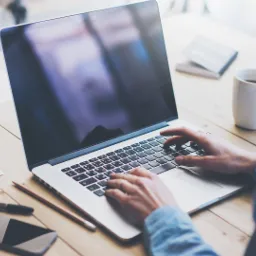 Osoba pisze na laptopie na drewnianym biurku, obok notatki, telefon, długopis, ołówek i kubek z napojem.