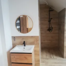 Nowoczesna łazienka z umywalką w szafce z drewnopodobnym fornirem, okrągłym lustrem z metalową ramą i kabiną prysznicową wyłożoną płytkami imitującymi drewno, z czarną armaturą.