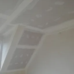 Skośny sufit z płyt kartonowo-gipsowych, częściowo zaszpachlowany, z widocznymi łączeniami i narożnikami, okno w tle.