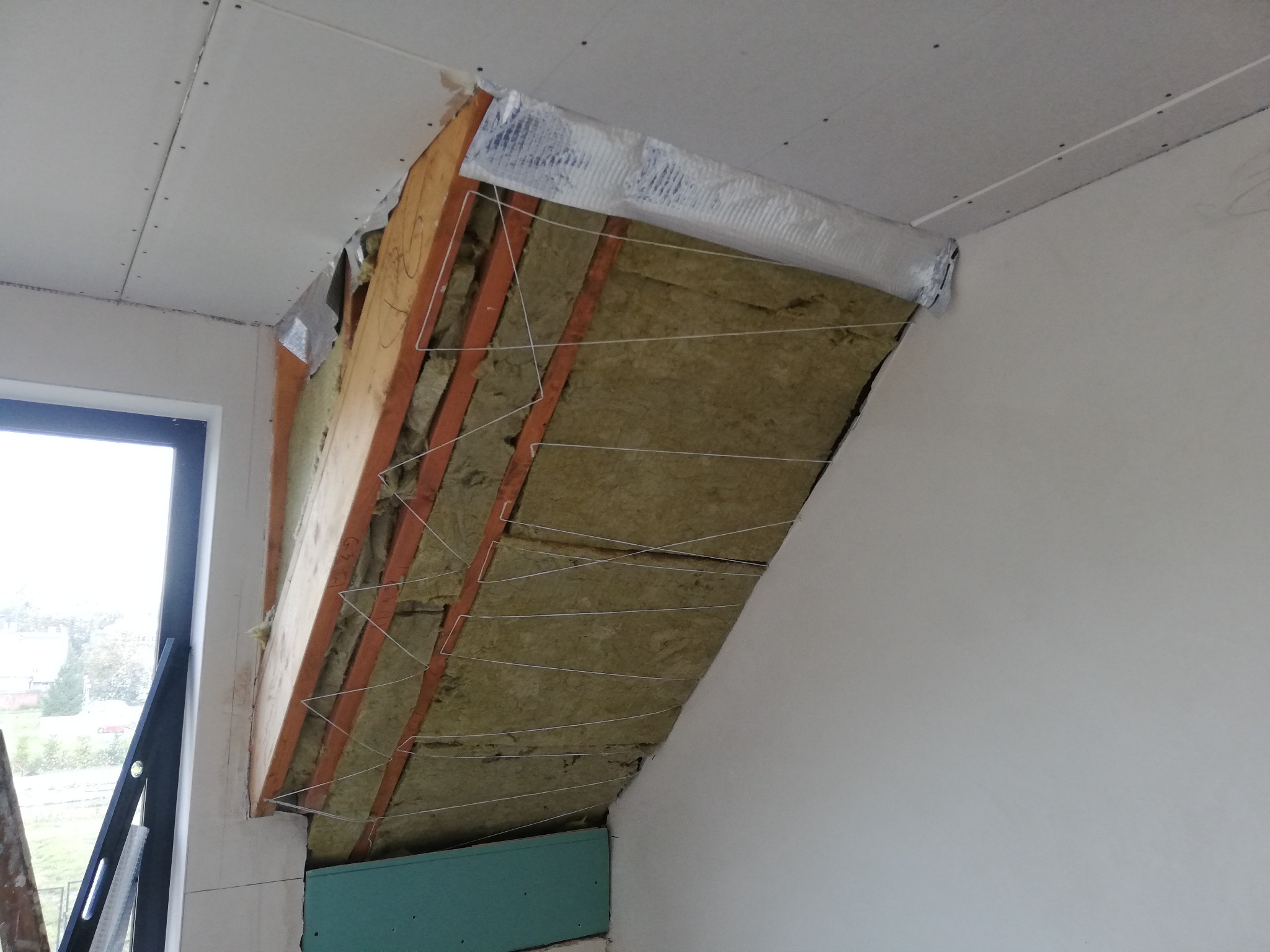 Skośna ściana z odsłoniętą konstrukcją drewnianą, izolacją z wełny mineralnej i częściowo zamontowaną płytą kartonowo-gipsową, widoczny poziomica przy oknie.