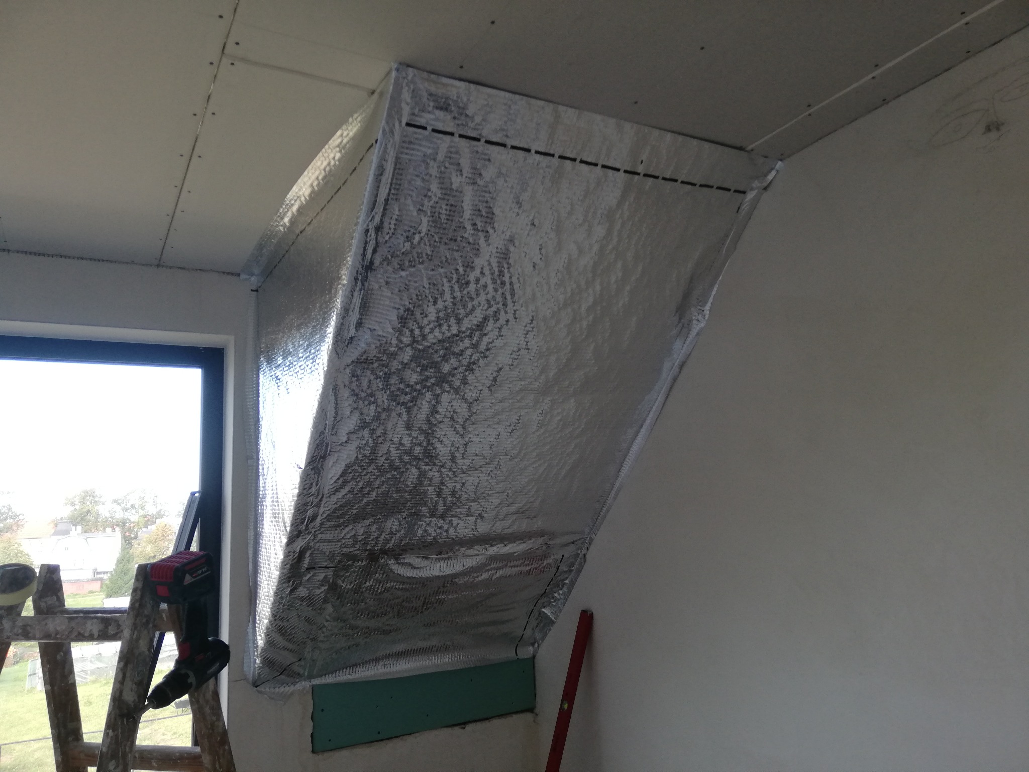 Skośne poddasze z izolacją termiczną w folii aluminiowej, częściowo obłożone płytą gipsowo-kartonową, drabina malarska z wkrętarką w tle okna.