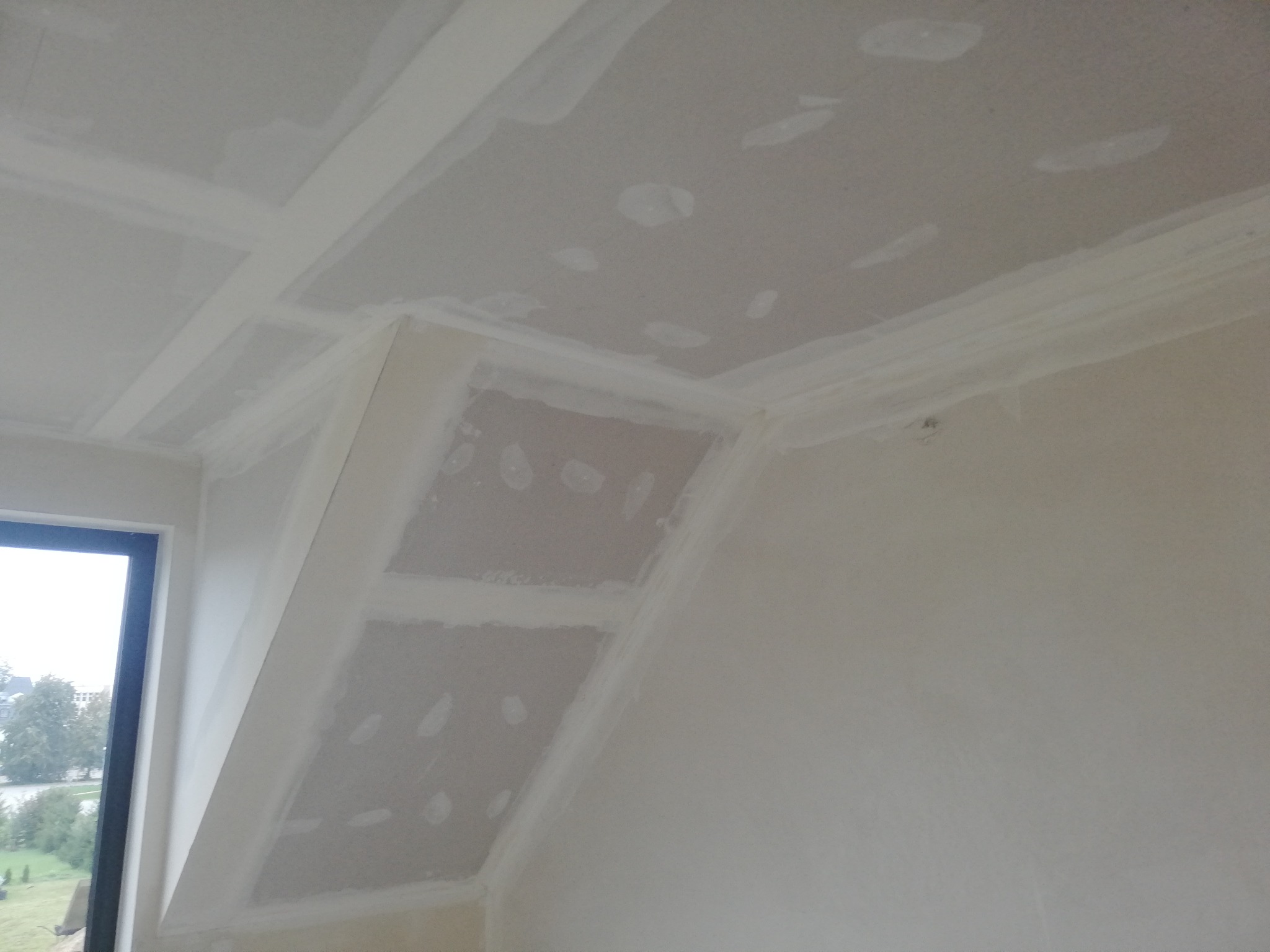 Skośny sufit z płyt kartonowo-gipsowych, częściowo zaszpachlowany, z widocznymi łączeniami i narożnikami, okno w tle.