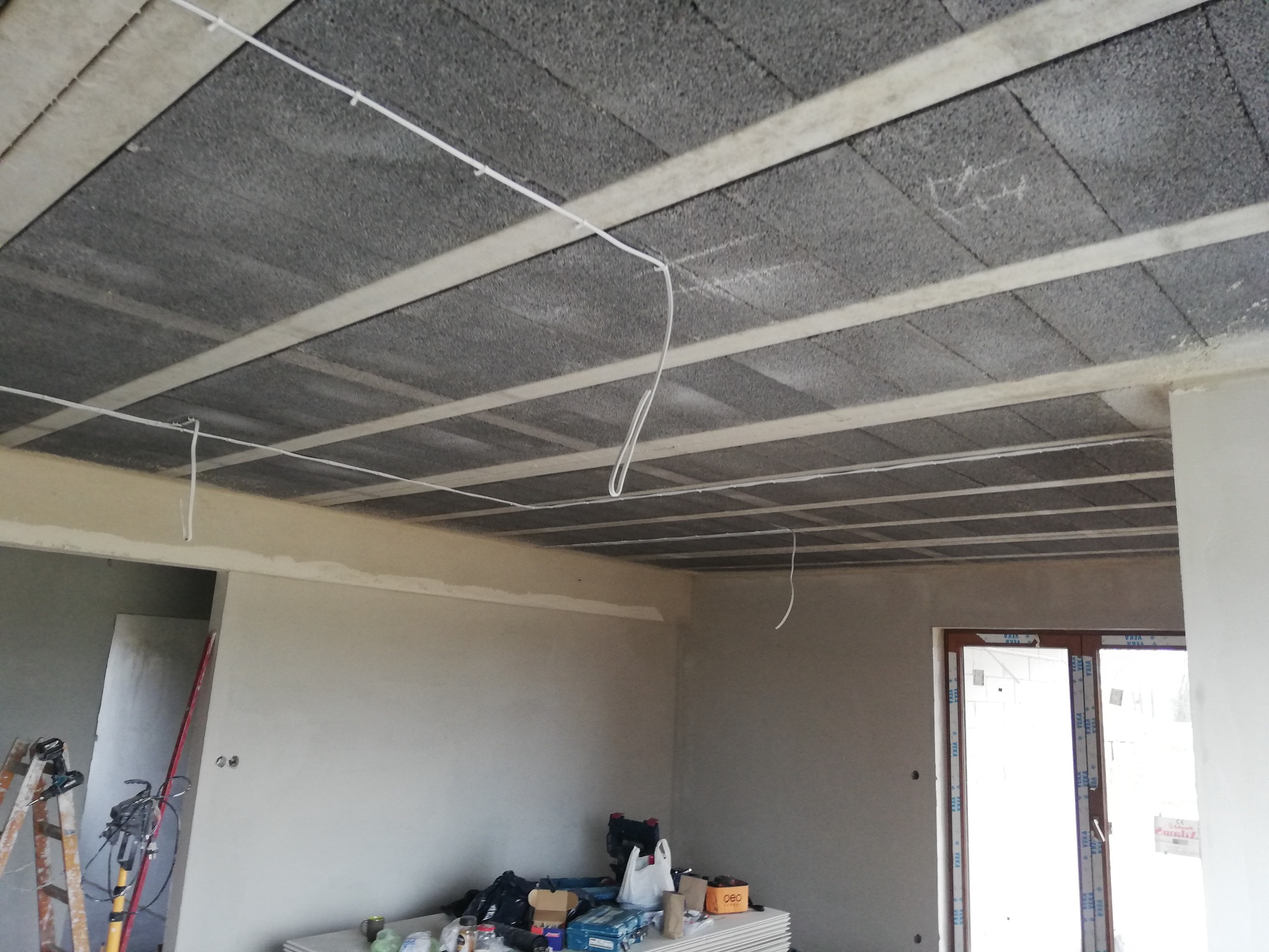 Surowy strop z betonowych pustaków i białych belek, widoczne przewody elektryczne, ściany w szarym tynku, narzędzia i materiały budowlane na podłodze, okno balkonowe.