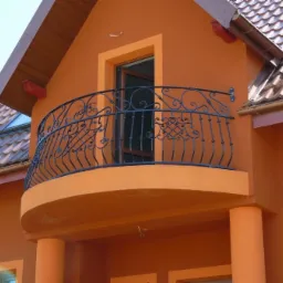 Pomarańczowy budynek z balkonem z kutą, czarną balustradą o ozdobnym wzorze, widok na brązowy dach z lukarną.