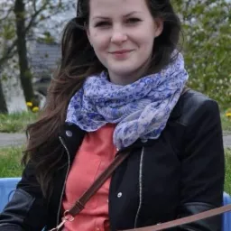 Portret młodej kobiety w czarnej kurtce, z niebieskim szalem w kwiaty i okularami przeciwsłonecznymi na głowie, siedzącej na niebieskim krześle na zewnątrz w słoneczny dzień.