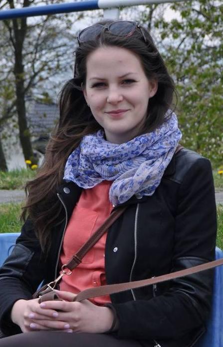 Portret młodej kobiety w czarnej kurtce, z niebieskim szalem w kwiaty i okularami przeciwsłonecznymi na głowie, siedzącej na niebieskim krześle na zewnątrz w słoneczny dzień.