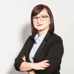 Portret kobiety w czarnym żakiecie i okularach, z rękami skrzyżowanymi na piersiach, na jasnym tle.