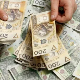 Stos banknotów 200 i 100 złotych w dłoniach, rozrzucone na jasnym tle, z widocznymi numerami seryjnymi i portretami.