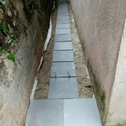 Wąska ścieżka z szarych płyt chodnikowych układana wzdłuż ściany budynku, widoczne wystające pręty stabilizujące, podłoże z piasku i geowłókniny.