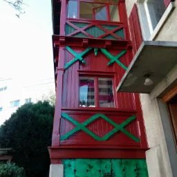 Czerwony, drewniany balkon z zielonymi zdobieniami w kształcie krzyży, widoczny z dołu, z otwartym oknem na górze i zielonymi, pionowymi deskami na dole.