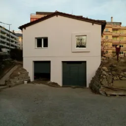 Dwukondygnacyjny dom z dwoma garażami, świeżo pomalowaną fasadą w kolorze jasnego beżu i ciemnozielonymi drzwiami garażowymi, widziany z poziomu podjazdu.