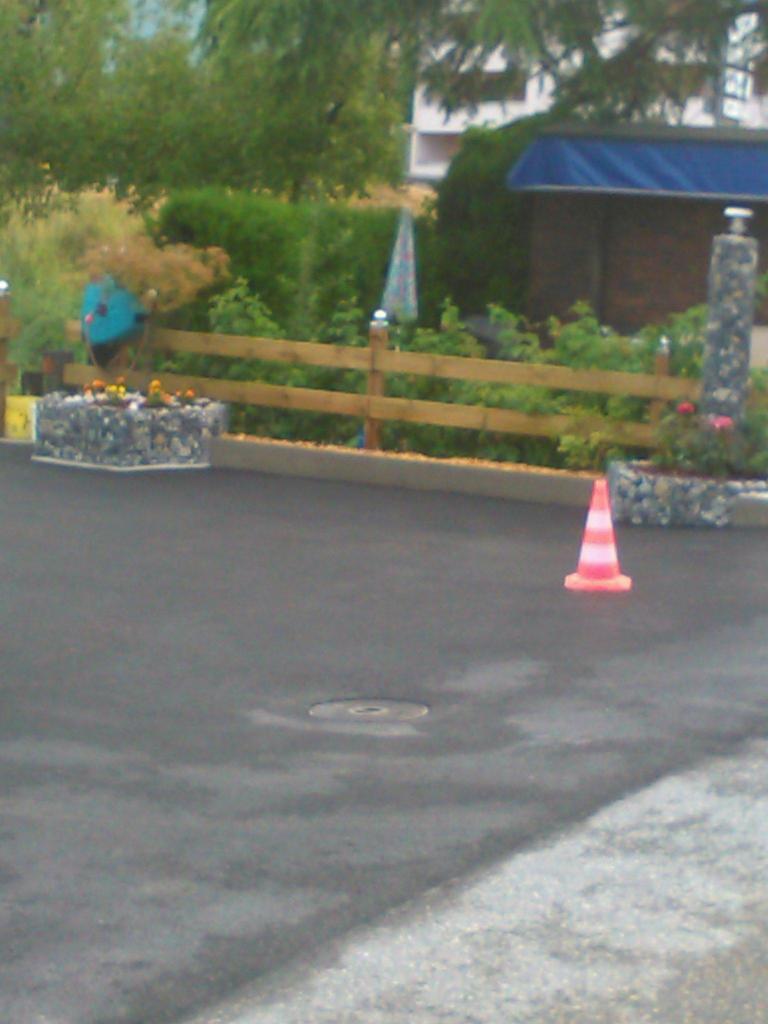 Gabionowe donice z kwiatami i drewniane ogrodzenie z oświetleniem, widoczne na tle zieleni, na pierwszym planie stożek drogowy na asfaltowej nawierzchni.