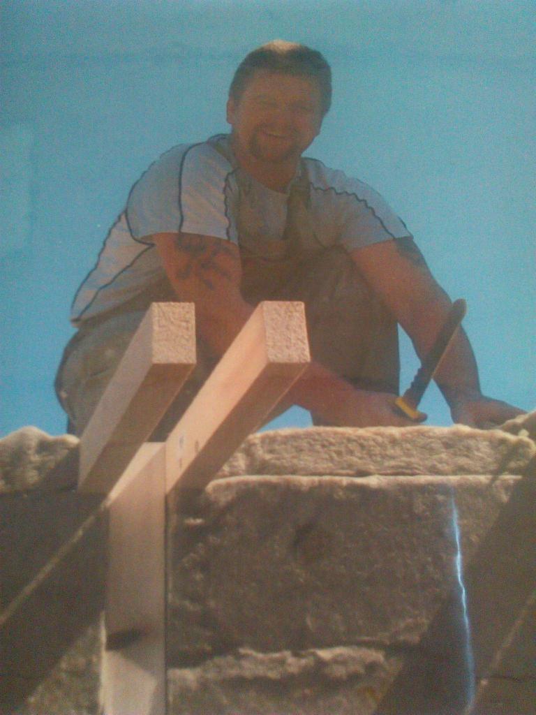 Mężczyzna z tatuażem na ramieniu, uśmiechnięty, w trakcie montażu drewnianej konstrukcji na ocieplonym balkonie, trzymając w ręku nóż do wełny mineralnej.