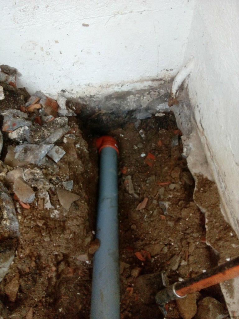 Szary przewód kanalizacyjny z pomarańczowym łącznikiem w wykopie przy ścianie, gruz i ziemia.