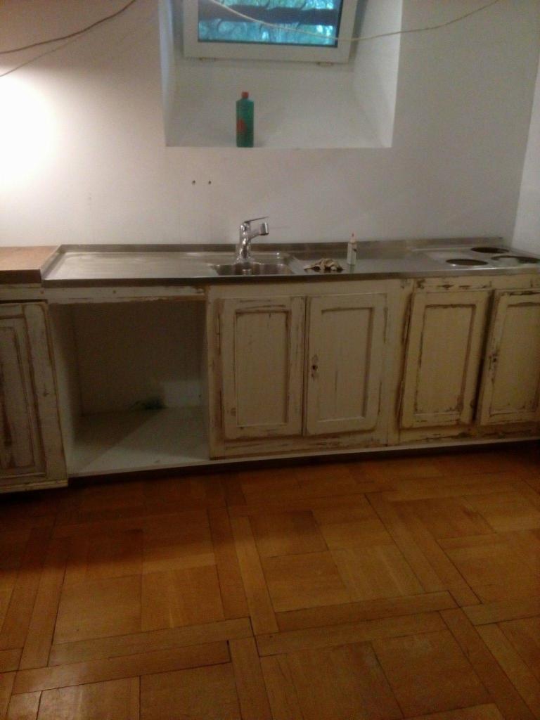 Fragment kuchni z drewnianą podłogą parkietową, zabudowa kuchenna w stylu rustykalnym z białymi, przecieranymi frontami, metalowy blat z wbudowanym zlewem i płytą grzewczą, na ścianie małe okno...