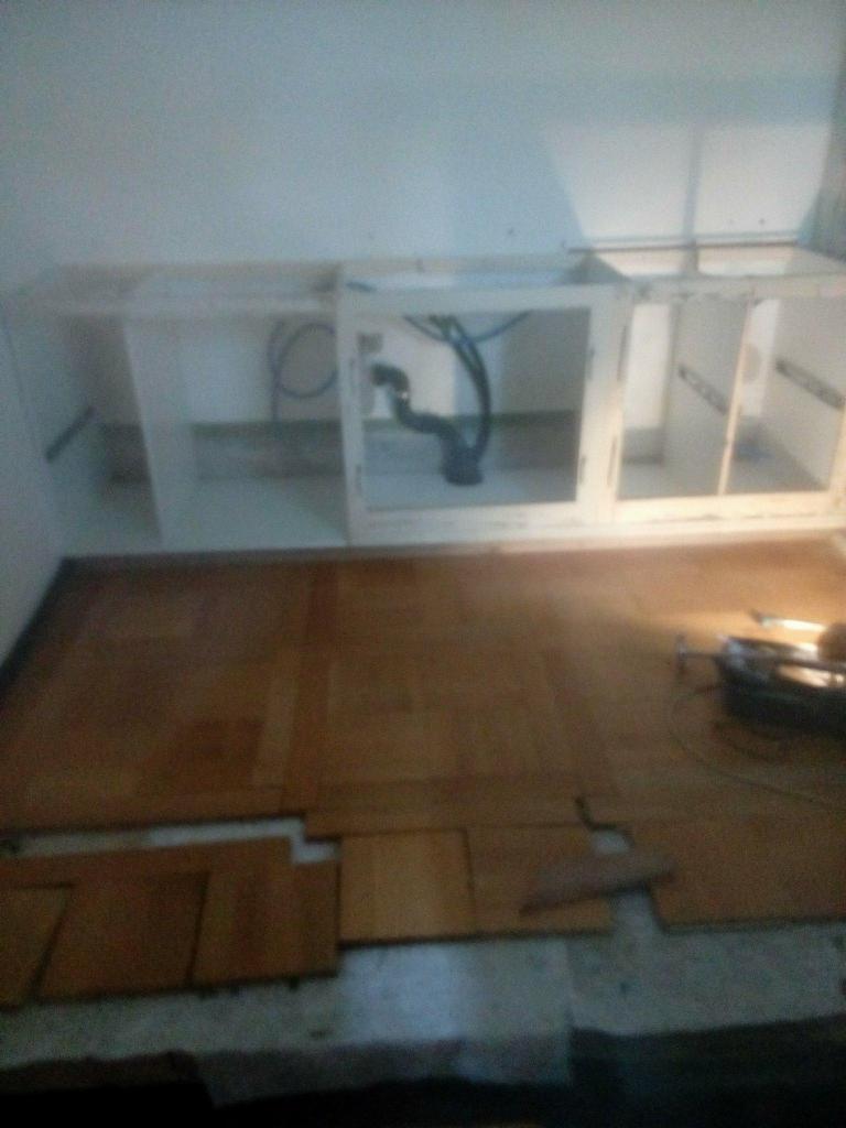 Montaż drewnianej podłogi i szafek kuchennych pod zlewem, widoczne rury odpływowe i narzędzia na podłodze.