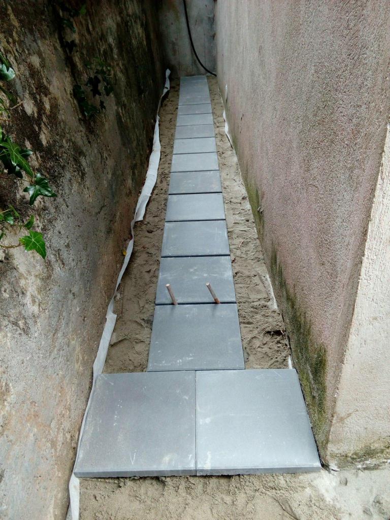 Wąska ścieżka z szarych płyt chodnikowych układana wzdłuż ściany budynku, widoczne wystające pręty stabilizujące, podłoże z piasku i geowłókniny.