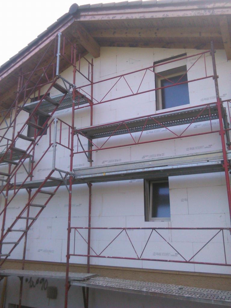 Dom w trakcie ocieplania styropianem, widoczne rusztowanie z metalowymi schodami i dwa okna.