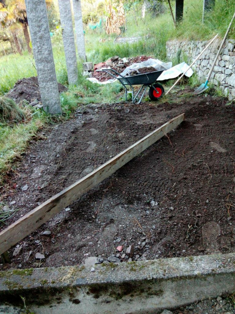 Przygotowanie terenu pod ogród: wyrównana ziemia z deska, taczka z kompostem, łopata oparta o kamienny murek w tle, wczesna faza prac.