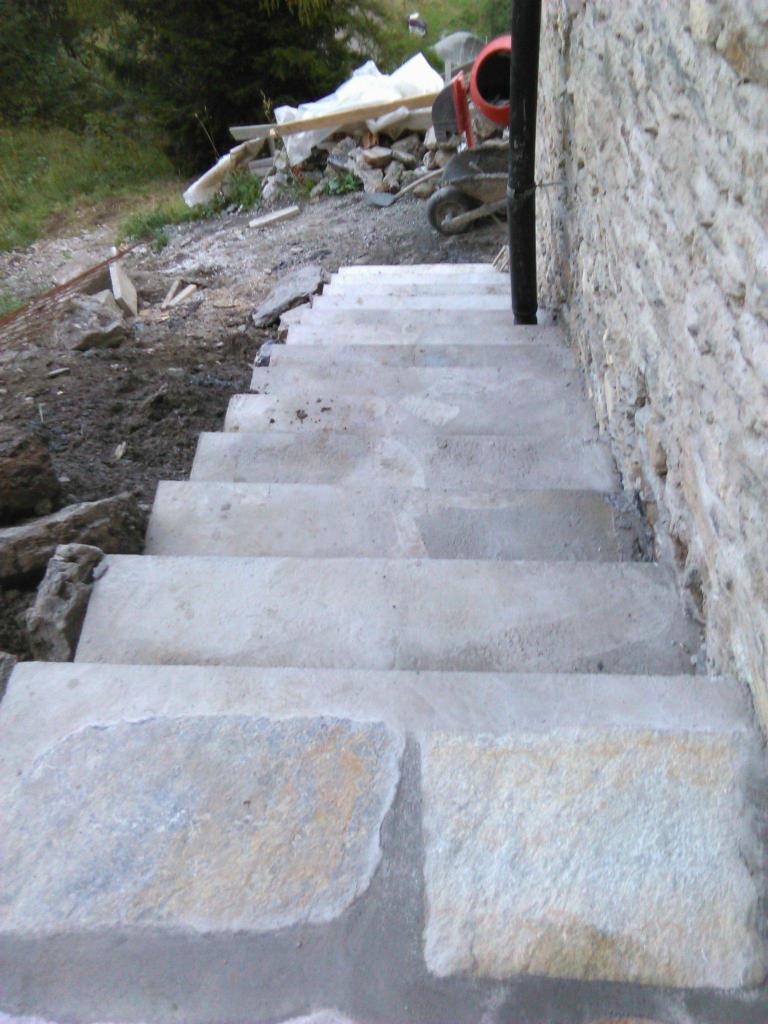 Kamienne schody prowadzące w górę, z widocznym betonem między stopniami i ścianą z kamienia po prawej stronie, w tle gruz, taczka i betoniarka.