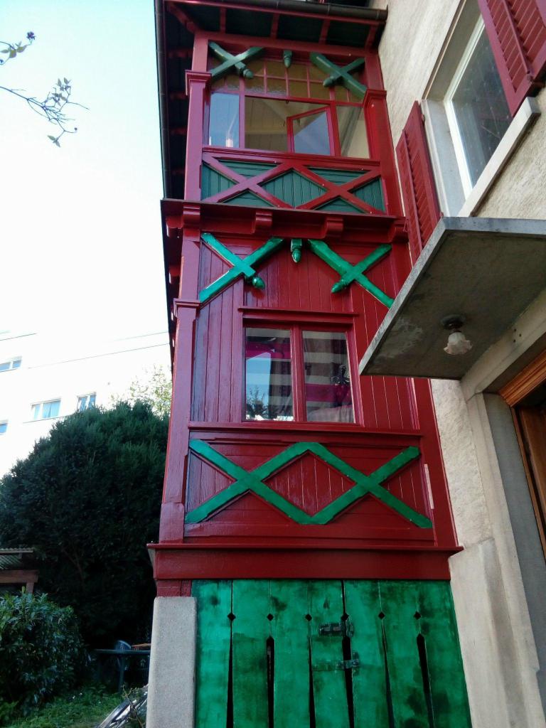 Czerwony, drewniany balkon z zielonymi zdobieniami w kształcie krzyży, widoczny z dołu, z otwartym oknem na górze i zielonymi, pionowymi deskami na dole.