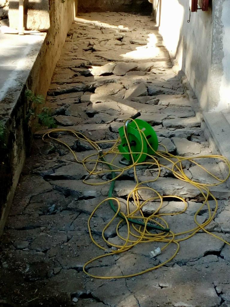 Zniszczony chodnik z popękaną nawierzchnią i leżącym na nim zwiniętym, żółtym przedłużaczem w zielonej obudowie.