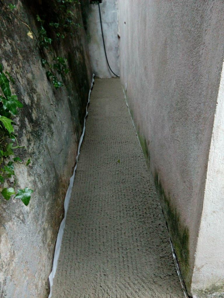 Wąskie przejście między dwiema ścianami po świeżo wylanej wylewce betonowej, zabezpieczonej folią przy ścianie z roślinnością.