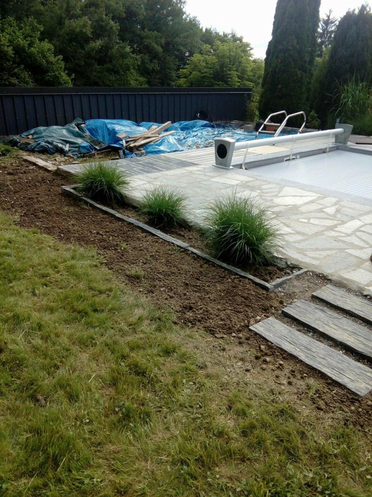 Fragment ogrodu z basenem w trakcie prac: świeżo przekopana ziemia, ozdobne trawy, kamienna ścieżka prowadząca do zadaszonego basenu, w tle składowane materiały budowlane pod plandeką.