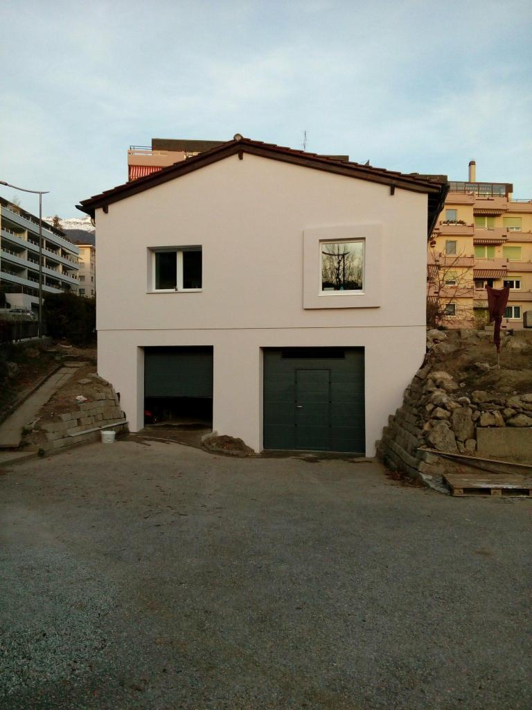 Dwukondygnacyjny dom z dwoma garażami, świeżo pomalowaną fasadą w kolorze jasnego beżu i ciemnozielonymi drzwiami garażowymi, widziany z poziomu podjazdu.