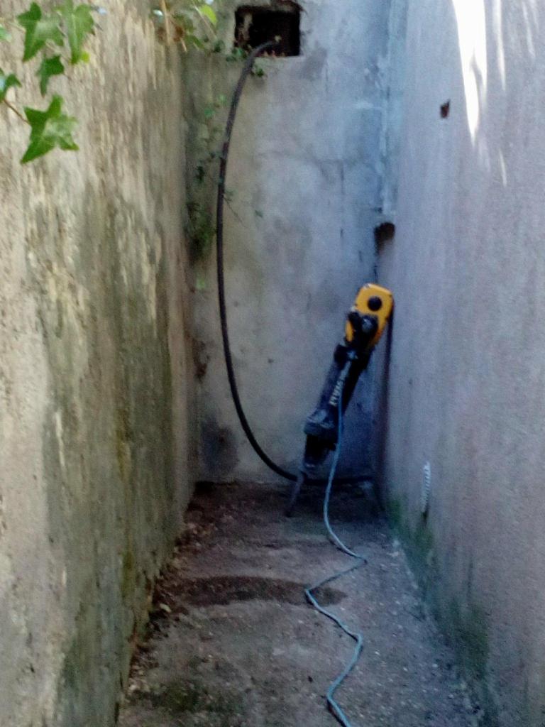 Wąski, zaniedbany korytarz z betonowymi ścianami, gdzie stoi młot udarowy z żółtą obudową, podłączony do czarnego węża, prowadzącego do otworu w górnej części ściany.