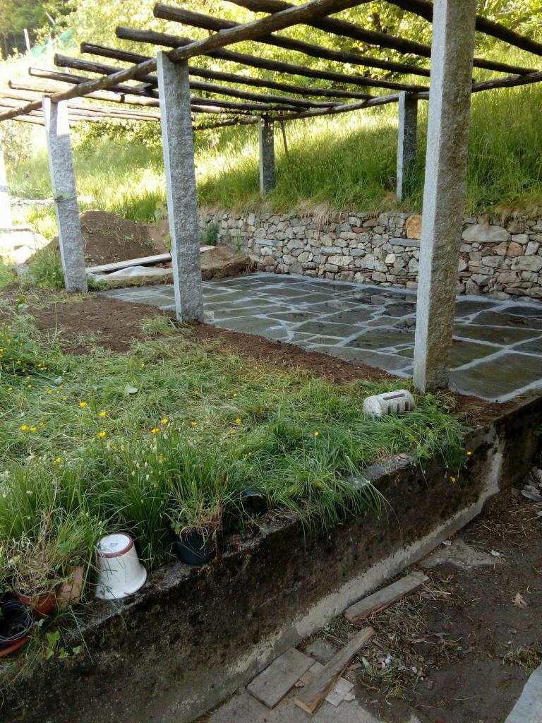 Pergola z kamiennymi słupami i nieregularną kamienną posadzką w ogrodzie, widok na kamienny murek i zbocze porośnięte trawą, doniczki z roślinami w pierwszym planie.